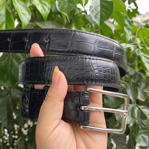 Puede incluir: Un cinturón negro con un patrón texturizado de relieve de cocodrilo y una hebilla de metal plateado pulido. La correa del cinturón tiene costuras visibles a lo largo de los bordes y una serie de agujeros perforados para el ajuste.