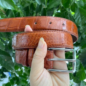 Puede incluir: Un cinturón marrón con un patrón texturizado de piel de cocodrilo, que presenta una hebilla de metal plateado pulido y costuras marrones a juego en los bordes. El cinturón tiene varios agujeros preperforados y un forro interior liso y oscuro.