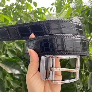 Puede incluir: Un cinturón negro con un patrón de piel de cocodrilo brillante y texturizado con un detalle de borde trenzado. El cinturón está equipado con una hebilla de metal plateado pulido que tiene un marco rectangular y un mecanismo de hebijón.