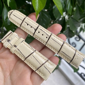 May include: A two-piece watch strap in a cream-colored leather with a glossy finish and a textured alligator-style pattern. The strap features dark brown stitching along the edges and includes a matching leather loop and buckle holes.