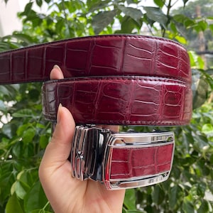 Puede incluir: Un cinturón de color burdeos intenso con una textura brillante de piel de cocodrilo. El cinturón incluye una hebilla metálica rectangular con una incrustación de piel de cocodrilo burdeos a juego en el centro y bordes de metal plateado pulido.