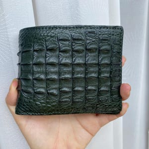 May include: A dark green bifold wallet featuring a textured, raised scale pattern across the exterior surface. The wallet has a rectangular shape with visible stitching along the edges and a distinct, segmented crocodile-like skin appearance.