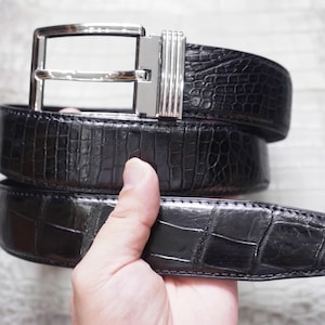 May include: A black belt with a textured, embossed pattern resembling reptile skin. The belt features a polished, rectangular silver-tone metal buckle with a vertical ribbed detail on the side, secured by a single prong and a matching metal loop.