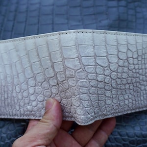 May include: A bi-fold wallet crafted from off-white leather featuring a prominent, raised crocodile skin texture. The surface displays a detailed scale pattern with subtle shading, and the edges are finished with clean, matching stitching.