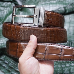 Puede incluir: Un cinturón marrón con un patrón texturizado de piel de cocodrilo brillante y una hebilla de metal plateado pulido. El cinturón presenta una hebilla rectangular con un perno y un detalle de presilla de metal estriado en tono marrón.