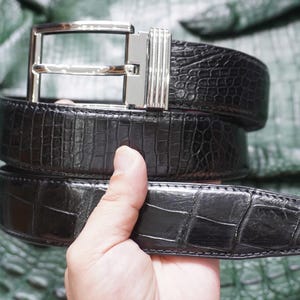 Puede incluir: Un cinturón negro con un patrón texturizado en relieve que recuerda a la piel de reptil. El cinturón cuenta con una hebilla de metal plateado pulido con un marco rectangular y un detalle lateral acanalado. La correa tiene un acabado brillante.