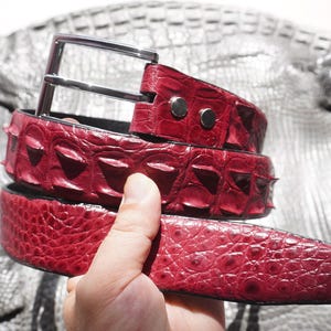 Puede incluir: Un cinturón de color rojo intenso con un patrón de escamas texturizado en relieve y una hebilla de metal plateado pulido. La correa del cinturón muestra una textura superficial irregular y distintiva, característica del cuero exótico, asegurada con dos remaches de metal plateado.