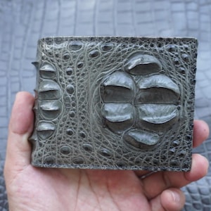 Peut inclure: Un portefeuille pliable en cuir gris texturé présentant un motif d'écailles en relief. La surface affiche une disposition distincte de grandes écailles arrondies et d'écailles plus fines, créant un aspect tactile et multidimensionnel.