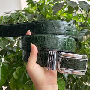 May include: A dark green belt featuring a textured crocodile skin pattern and a polished silver-tone metal buckle. The belt strap has a glossy finish and is coiled, showcasing the detailed embossed scales across the entire length of the leather.