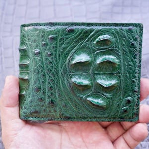May include: A bifold wallet crafted from deep emerald green leather with a prominent, raised crocodile skin texture. The surface features large, distinct scales and a glossy finish, showcasing the natural pattern of the hide across the entire exterior.