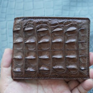 May include: A brown bifold wallet featuring a textured, raised scale pattern across the exterior surface. The wallet has a rectangular shape with visible stitching along the edges, showcasing a distinct, repeating organic pattern on the front panel.