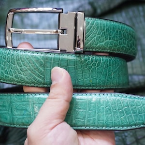 Puede incluir: Un cinturón de color verde brillante con un patrón texturizado y en relieve que imita la apariencia de la piel de reptil. El cinturón está equipado con una hebilla de metal plateado pulido que tiene un marco rectangular y un perno.