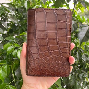 Può includere: Un portafoglio rettangolare in pelle marrone caratterizzato da una texture in rilievo che imita la pelle di coccodrillo. Il portafoglio presenta una finitura lucida e cuciture visibili lungo i bordi, con un design strutturato a orientamento verticale.