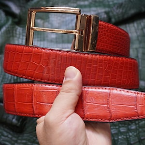Op de afbeelding: Een felrode riem met een gestructureerd krokodillenleerpatroon en een gepolijste goudkleurige metalen gesp. De riemband vertoont over de gehele lengte een consistent rechthoekig schubbenpatroon, aangevuld met een metalen framegesp.