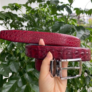 Op de afbeelding: Een dieprode riem met een textuur van krokodillenleer en een gepolijste zilverkleurige metalen gesp. De riemband vertoont over de gehele lengte een consistente schubachtige oppervlakteafwerking, gecombineerd met een rechthoekige metalen gesp.