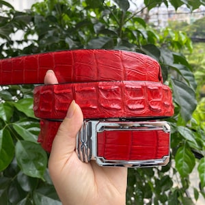 Op de afbeelding: Een felrode riem met een gestructureerd, reliëfpatroon dat het uiterlijk van reptielenhuid nabootst. De riem is voorzien van een rechthoekige, zilverkleurige metalen gesp met een gepolijste afwerking en een decoratief element aan de zijkant.