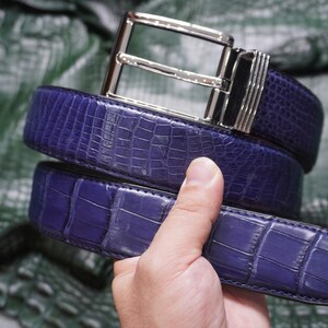 May include: A deep purple belt featuring a textured, embossed crocodile skin pattern and a polished silver-tone metal buckle with a rectangular frame and a multi-ribbed side detail, showcasing a distinct scale-like surface finish throughout.