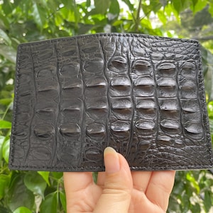Puede incluir: Una cartera rectangular de color negro que presenta un patrón de piel de cocodrilo texturizado, prominente y en relieve en toda la superficie. La cartera tiene costuras visibles a lo largo de los bordes y un acabado brillante que resalta los detalles.