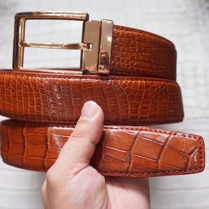 Puede incluir: Un cinturón marrón con textura de piel de cocodrilo brillante y una hebilla de metal dorado pulido. El cinturón presenta una hebilla de marco rectangular con un perno dorado a juego y un borde acabado liso a lo largo de la correa.