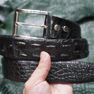 Op de afbeelding: Een zwarte riem met een textuur van krokodillenleer en een gepolijste zilverkleurige metalen gesp. De riem heeft een opvallend verhoogd schubbenpatroon over de hele lengte en is vastgezet met twee zilverkleurige metalen klinknagels bij de gesp.