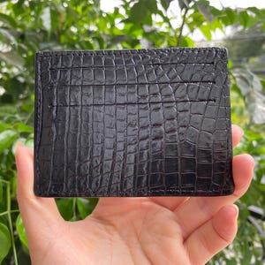 May include: A rectangular black card holder featuring a textured crocodile skin pattern. The front surface displays three horizontal slots for cards, with visible stitching along the edges and a glossy finish across the entire exterior surface.