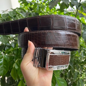 Puede incluir: Un cinturón marrón oscuro con textura de piel de cocodrilo brillante y una hebilla rectangular de metal plateado. La hebilla presenta una incrustación central de material con textura de cocodrilo a juego y un marco metálico pulido.