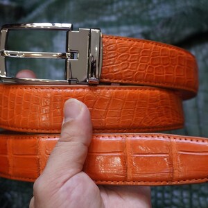 Puede incluir: Un cinturón de color naranja brillante con un patrón texturizado de piel de cocodrilo y una hebilla de metal plateado pulido. La correa del cinturón tiene costuras visibles en los bordes y está enrollada para mostrar el detalle en relieve.