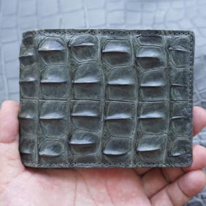 Puede incluir: Una cartera plegable de cuero texturizado gris oscuro que presenta un patrón de escamas en relieve en toda la superficie exterior. La cartera tiene una forma rectangular con costuras visibles a lo largo de los bordes y un acabado táctil distintivo.