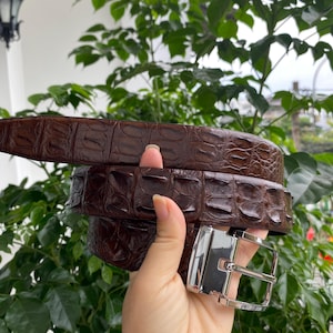 May include: A dark brown belt featuring a textured, embossed pattern resembling alligator skin. The belt is equipped with a polished, rectangular silver-tone metal buckle that has a clean, reflective finish and a standard prong mechanism.
