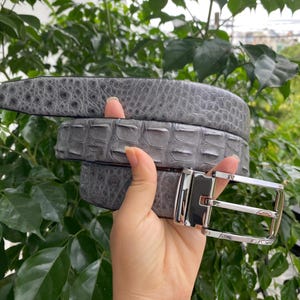 Puede incluir: Un cinturón gris con un patrón texturizado en relieve que imita la apariencia de piel exótica. El cinturón está equipado con una hebilla rectangular de metal plateado pulido y un perno de metal a juego para un cierre seguro.