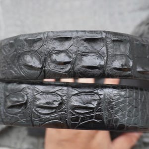 May include: A black belt featuring a prominent, raised, and textured scale pattern characteristic of crocodile skin. The surface has a glossy, reflective finish with distinct, large, rectangular scales running along the length of the strap.