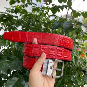 Op de afbeelding: Een felrode riem met een gestructureerd, in reliëf gemaakt krokodillenleerpatroon. De riem is voorzien van een gepolijste zilverkleurige metalen gesp met een rechthoekig frame en een centrale doorn, wat zorgt voor een veilige en verstelbare sluiting.