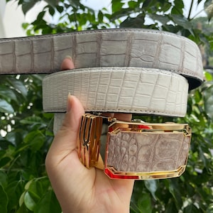 Op de afbeelding: Een riem met een gestructureerde, lichtgrijze en witte band met krokodillenpatroon. De riem heeft een rechthoekige goudkleurige metalen gesp met een bijpassende inleg met krokodillenpatroon in het midden en een gepolijste metalen rand.