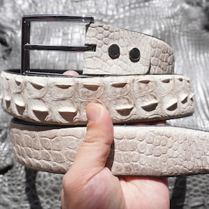 Op de afbeelding: Een crèmekleurige riem met een gestructureerd, verhoogd schubpatroon dat doet denken aan reptielenhuid. De riem is voorzien van een gepolijste gesp van donker metaal en twee ronde metalen klinknagels bij het bevestigingspunt.