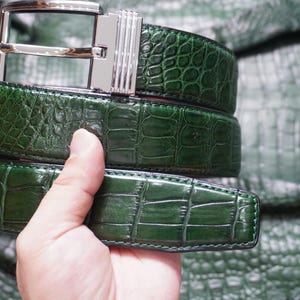 May include: A dark green belt with a glossy, textured crocodile-skin pattern and a polished silver-tone metal buckle. The belt features a rectangular buckle with vertical grooved detailing on the side and a smooth, shiny finish throughout.