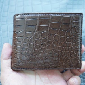 Op de afbeelding: Een donkerbruine bifold portemonnee met een glanzend, gestructureerd oppervlak en een opvallend reliëfpatroon dat het uiterlijk van krokodillenleer nabootst. De portemonnee heeft een rechthoekige vorm met zichtbare stiksels langs de randen.