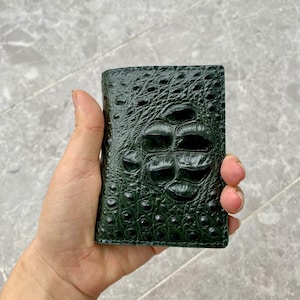 Può includere: Un portafoglio verde bosco scuro caratterizzato da un motivo in pelle di coccodrillo strutturato, prominente e in rilievo. La superficie presenta una finitura lucida con squame distinte e irregolari che variano di dimensioni sul corpo rettangolare.