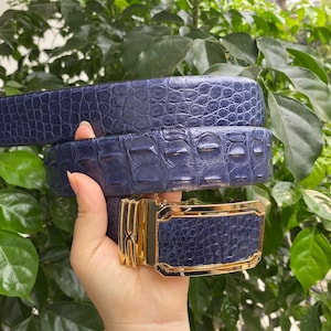 May include: A navy blue belt with a textured crocodile skin pattern and a rectangular gold-tone metal buckle. The belt features a repeating scale-like embossed design across the entire strap, complemented by a polished metallic buckle mechanism.
