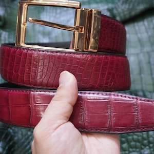 Puede incluir: Un cinturón de color rojo intenso con un patrón texturizado en relieve de piel de cocodrilo y una hebilla rectangular de metal pulido en tono dorado. El cinturón presenta un diseño clásico de hebilla de espiga y costuras visibles en los bordes.