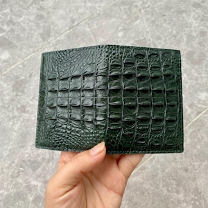 May include: A dark forest green bifold wallet featuring a prominent embossed crocodile skin texture. The exterior surface displays a repeating pattern of raised, rectangular scales that mimic the natural appearance of reptile hide.
