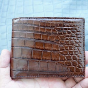 Peut inclure: Un portefeuille bifold marron doté d'une texture en relief façon peau de crocodile brillante. La surface présente un motif distinct de écailles rectangulaires répétitives sur tout l'extérieur, avec une finition polie qui reflète la lumière.