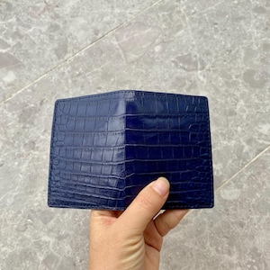 Può includere: Un portafoglio bifold blu scuro caratterizzato da una texture lucida in rilievo effetto pelle di coccodrillo. Il portafoglio ha una forma rettangolare con cuciture visibili lungo i bordi e una finitura liscia e lucida su tutta la superficie.
