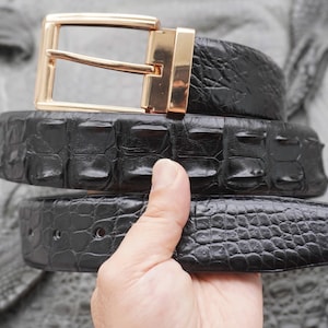 Puede incluir: Un cinturón negro con un patrón texturizado en relieve que recuerda a la piel de reptil. El cinturón cuenta con una hebilla rectangular de metal dorado pulido con un solo perno. La correa tiene un acabado brillante y costuras visibles.