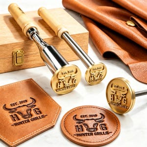 May include: Two metal branding irons with light wood handles and a separate circular branding head. The brass heads feature a bull head design with the text "EST. 2019" and "HUNTER GRILLS". Two leather pieces show the same bull logo embossed in the centre.