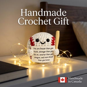 Könnte beinhalten: Ein weißer Häkel-Baseball mit roten Nähten, schwarzen Perlenaugen und einem kleinen Lächeln. Er hält einen hellen hölzernen Baseballschläger und ein weißes rechteckiges Schild mit dem Text: You are braver than you think, stronger than you let on, smarter than you imagine, and more loved than you know.