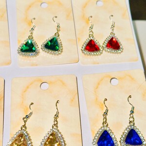 Peut inclure: Boucles d'oreilles pendantes avec des pendentifs triangulaires en pierres précieuses vertes, rouges, dorées et bleues. Chaque boucle d'oreille est sertie d'un métal argenté et entourée de petits cristaux clairs.