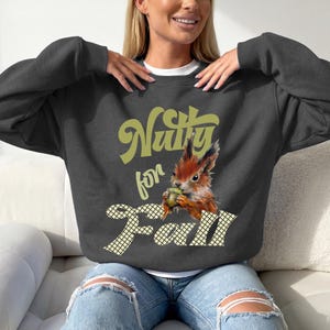 Könnte beinhalten: Ein anthrazitfarbenes Sweatshirt mit Rundhalsausschnitt und einer Grafik eines Eichhörnchens, das eine Eichel hält. Der Text "Nutty for Fall" ist in einer hellgelben Retro-Schrift gedruckt, wobei das Wort "Fall" auf einem weißen Gittermuster liegt.
