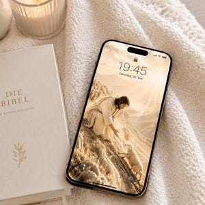 Könnte beinhalten: Ein Smartphone zeigt ein religiöses Bild von Jesus mit einem Lamm, das auf einer cremefarbenen Decke liegt. Daneben liegt ein Buch mit dem Titel "DIE BIBEL" und eine brennende Kerze in einem Glas. Die Szene ist in weiches, warmes Licht getaucht.