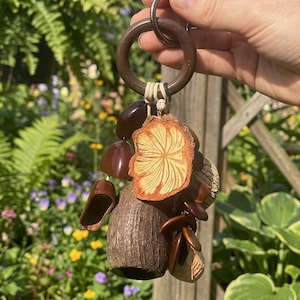 Puede incluir: Un llavero de madera con una colección de dijes en miniatura hechos a mano con forma de setas y bellotas. Los dijes son en tonos marrones y beige, con una seta grande y detallada en el centro. El llavero se sujeta con un anillo de madera.