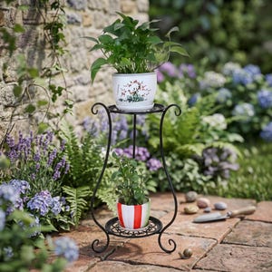 Op de afbeelding: Een zwartmetalen plantenstandaard met twee niveaus en decoratief krulwerk, met twee potplanten. De bovenste pot is wit met een bloemenmotief en de onderste pot is gestreept. De standaard staat op een stenen terras, omringd door groen.